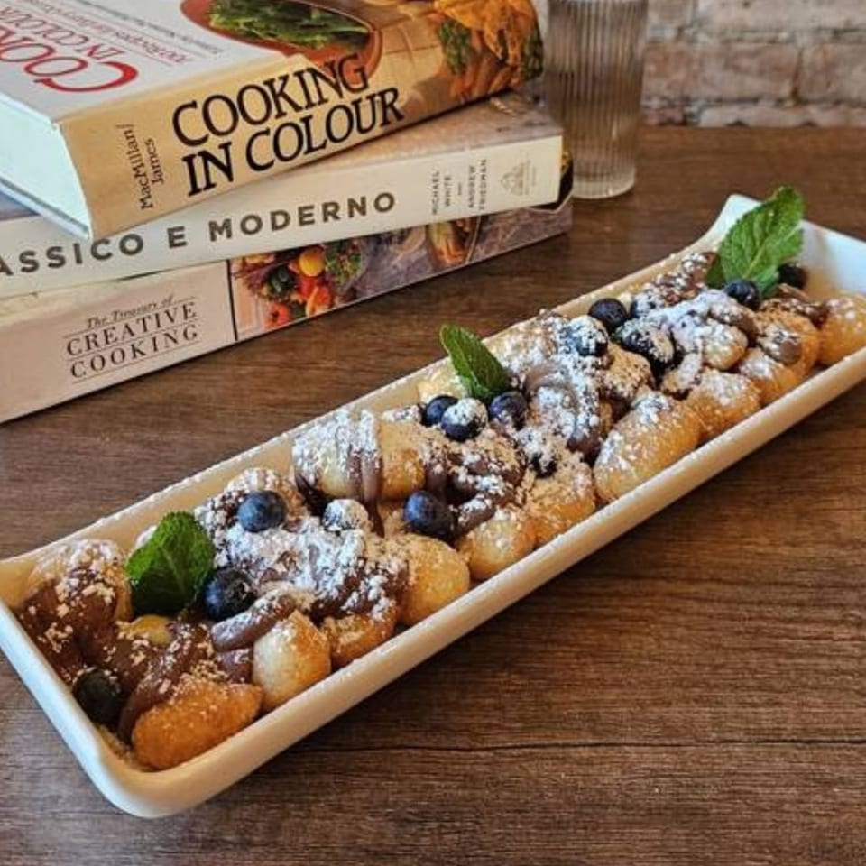 Assorted powdered-sugar pastries topped with blueberries and mint fill a rectangular platter beside Italian cooking books on a wooden table.