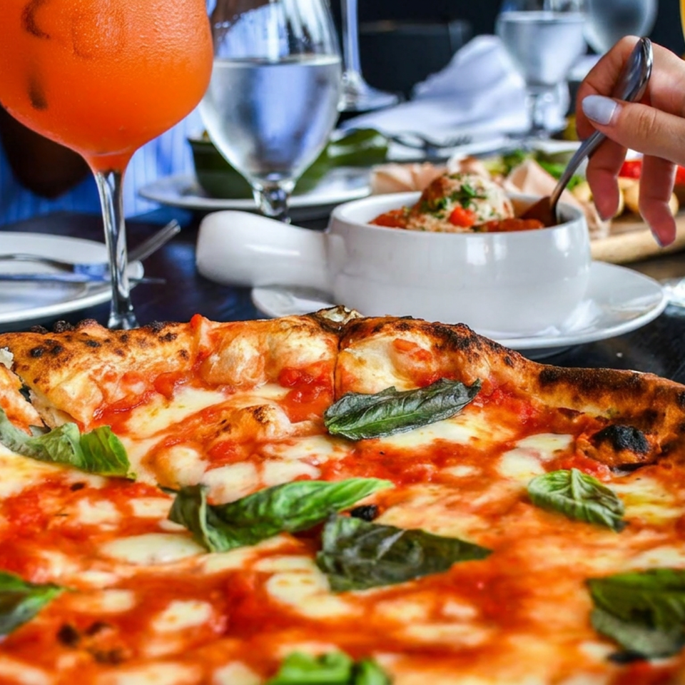 Neapolitan pizza topped with tomato, melted mozzarella, and fresh basil at a dining table with drinks and diners.