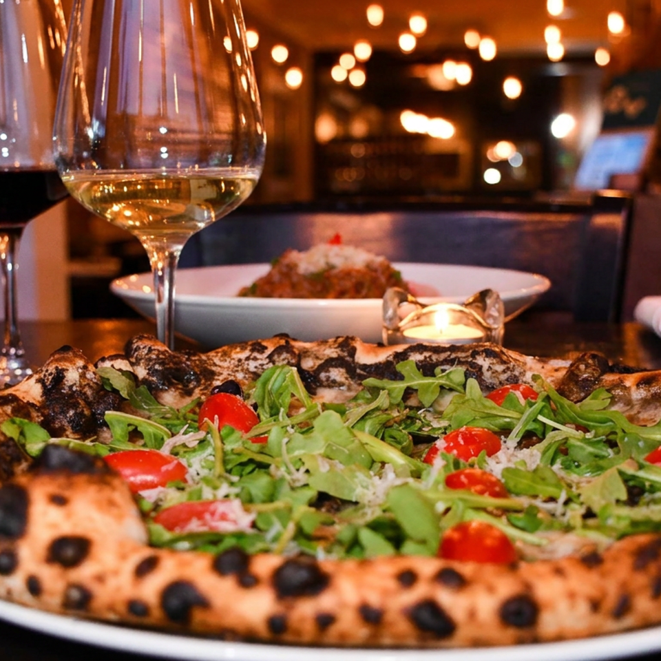 Wood-fired pizza topped with arugula, cherry tomatoes, and cheese beside red and white wine in a warmly lit restaurant.