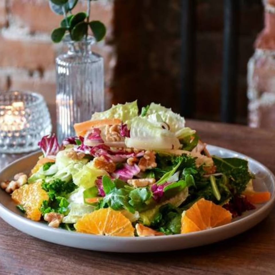 Colorful mixed green salad with orange slices, walnuts, vegetables, and edible flowers served on a plate in a cozy restaurant.