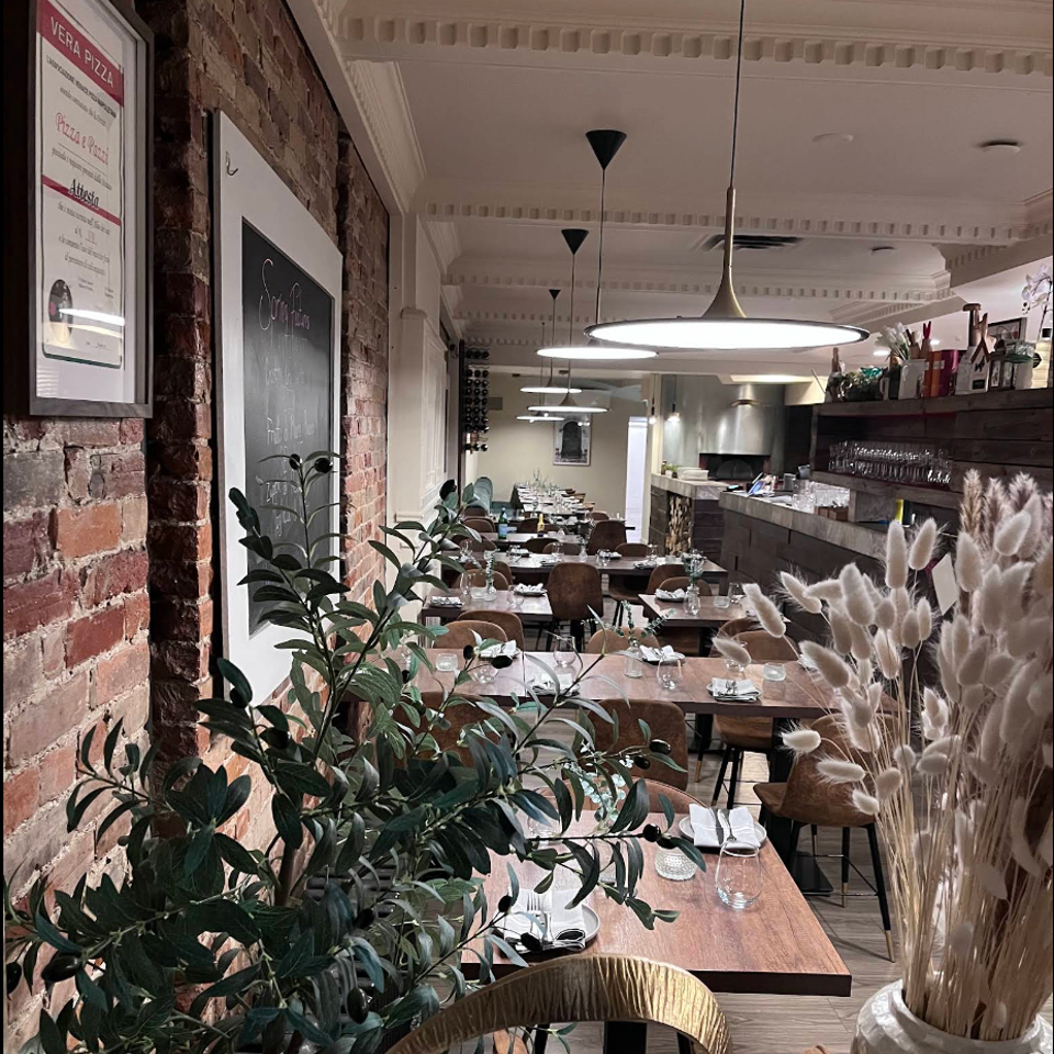 Warm Italian restaurant dining room with exposed brick, set tables, pendant lights, greenery, and a bar counter.