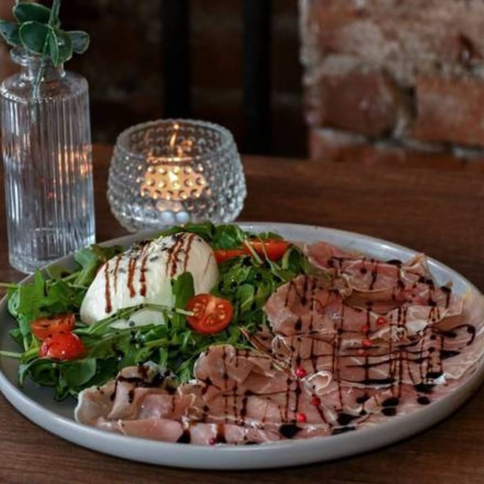 Platter of prosciutto, burrata, arugula, tomatoes, and balsamic glaze on a wooden restaurant table.