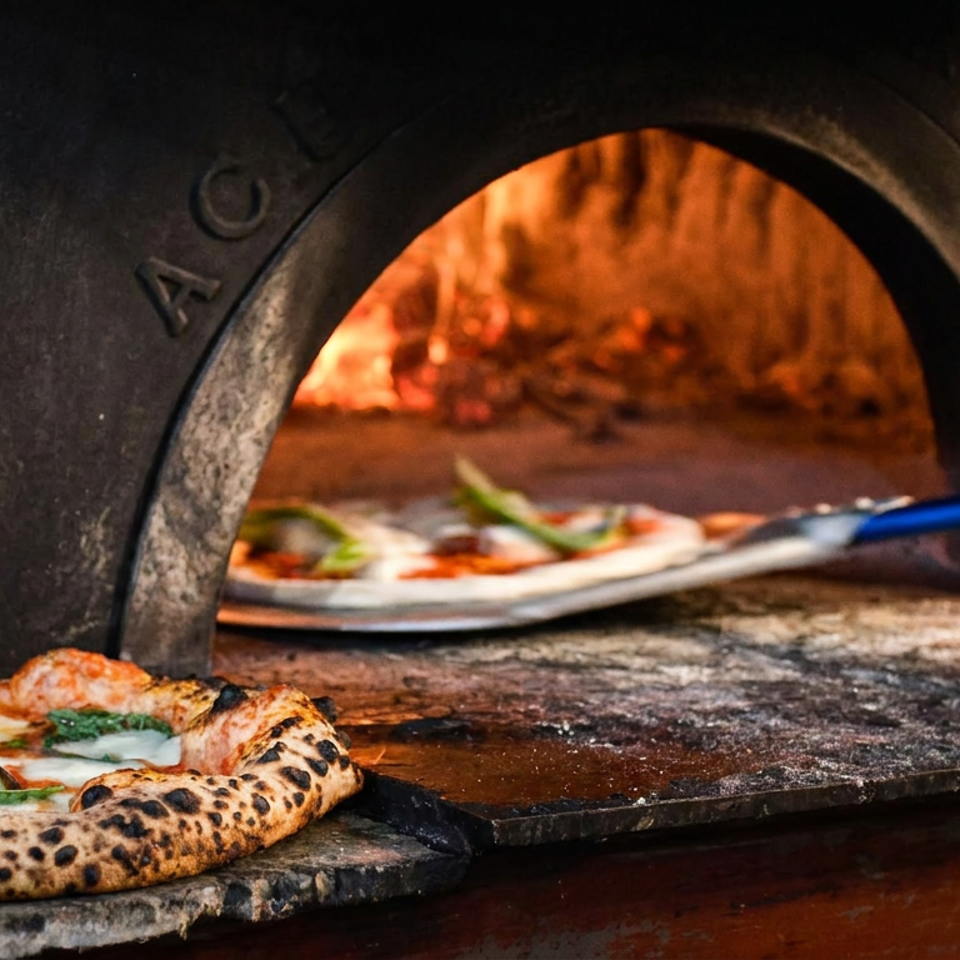 Wood-fired pizza with charred crust and basil sits beside a glowing brick oven as another pizza is prepared with a peel.